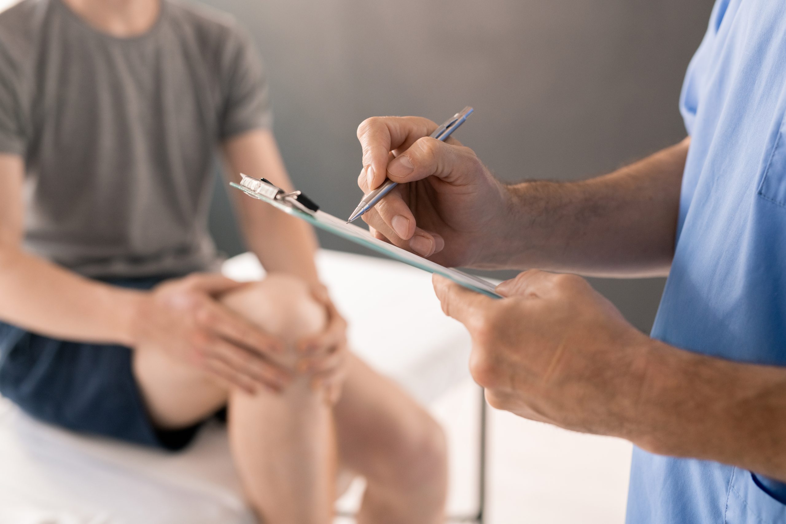 Hands of contemporary male physiotherapist with pen making prescriptions for patient with injured knee sitting on medical couch