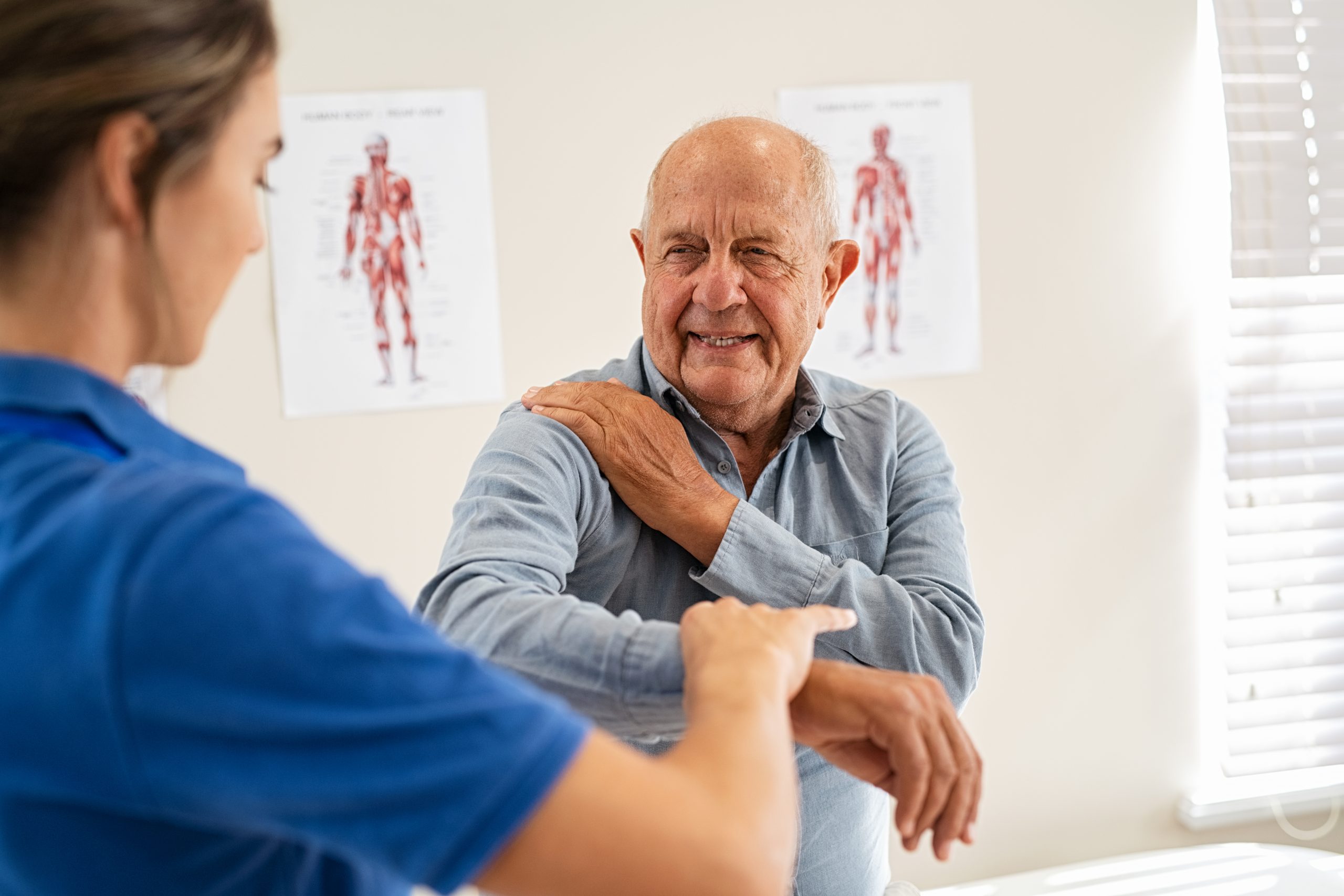 Young woman physiotherapist helping senior man with elbow exercise in clinic. Young woman doctor checking elbow of senior patient. Old man during an appointment with professional osteopath in private clinic working and massaging his shoulder pain.